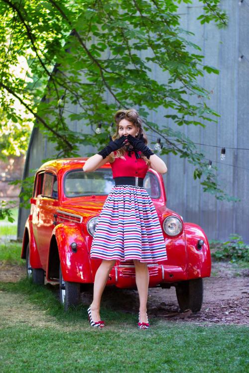 Дівчина у ретро сукні та чорних рукавичках, що стоїть перед ретро авто червоного кольору 