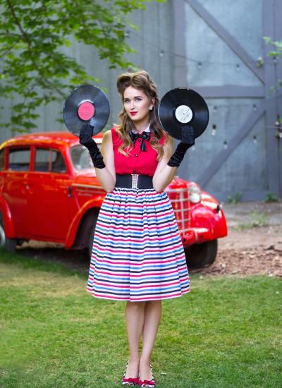 Стиляга у смужку Red & Blue & Black Дівчина в ретро-вбранні 50-х років тримає вінілові платівки на фоні червоного авто