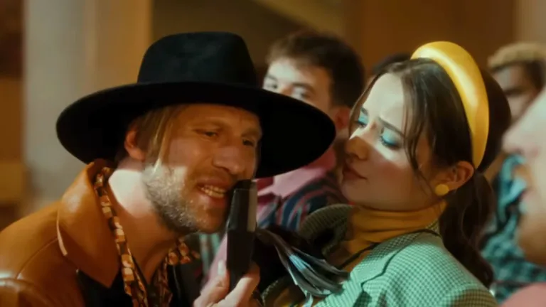 Іван Дорн у коричневій шкіряній куртці та капелюсі поруч із дівчиною в ретро-жакеті та жовтому обручі у кліпі «Мозоль»